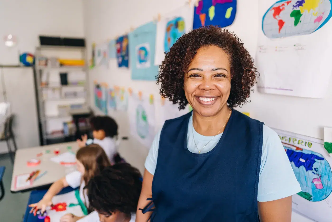 Professora sorrindo na sala de aula, com estudantes desenhando ao fundo