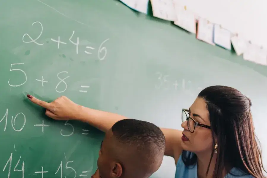 Professora aponta uma conta na lousa ao lado de um estudante.